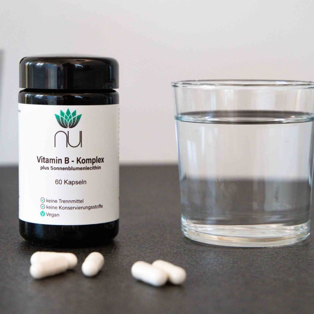 NUI Vitamin B-Komplex Glas steht neben einem Glas mit Wasser, daneben liegen Kapseln