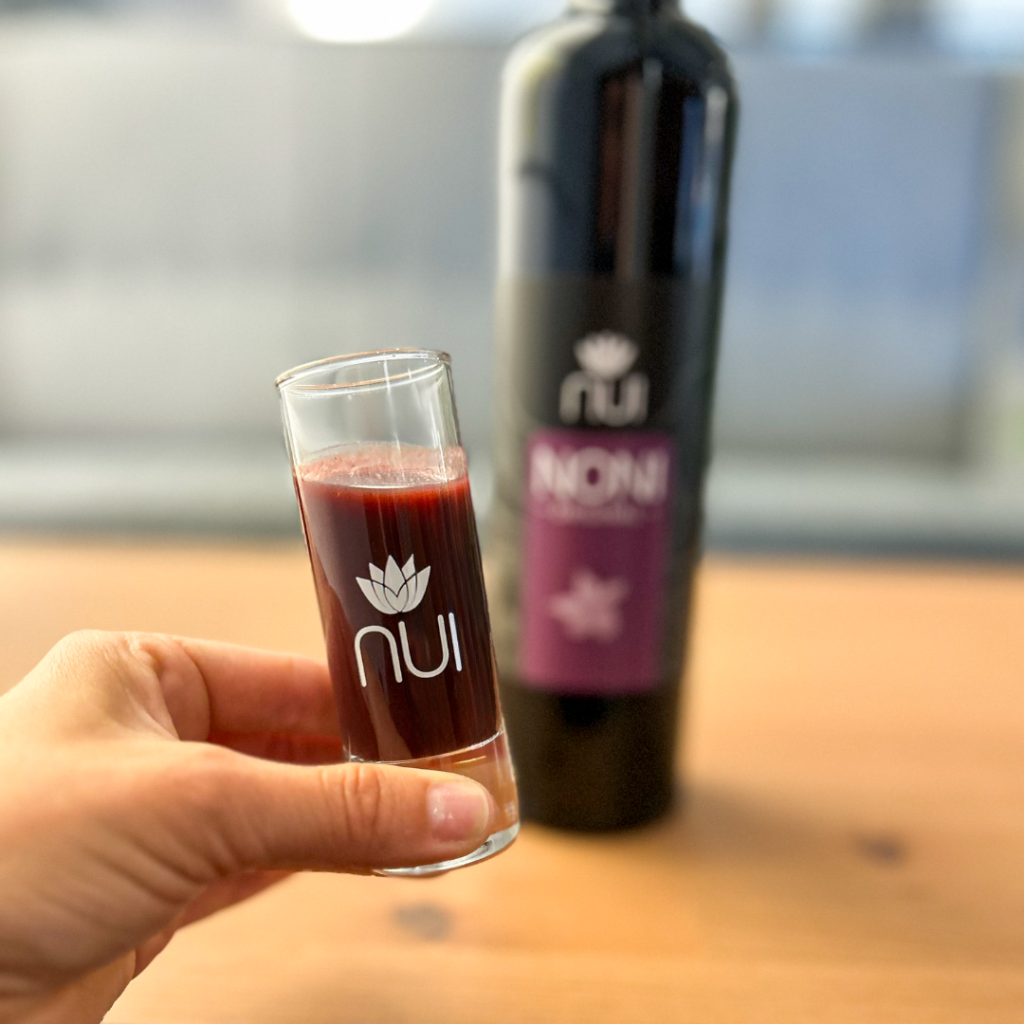 NUI Shotglas gefüllt mit Noni-Saft in der Hand im Hintergrund eine NUI NONI Original Flasche