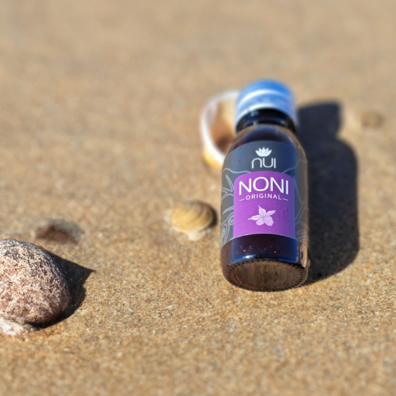 NUI NONI Shot Flasche liegt im Sand neben Muscheln und Steinen