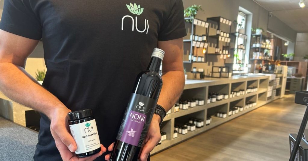 Mann mit schwarzem T-Shirt hat eine NUI Noni Flasche und ein Glas mit NUI Haut Haare Nägel in der Hand im Hintergrund der NUI Shop in Dornbirn