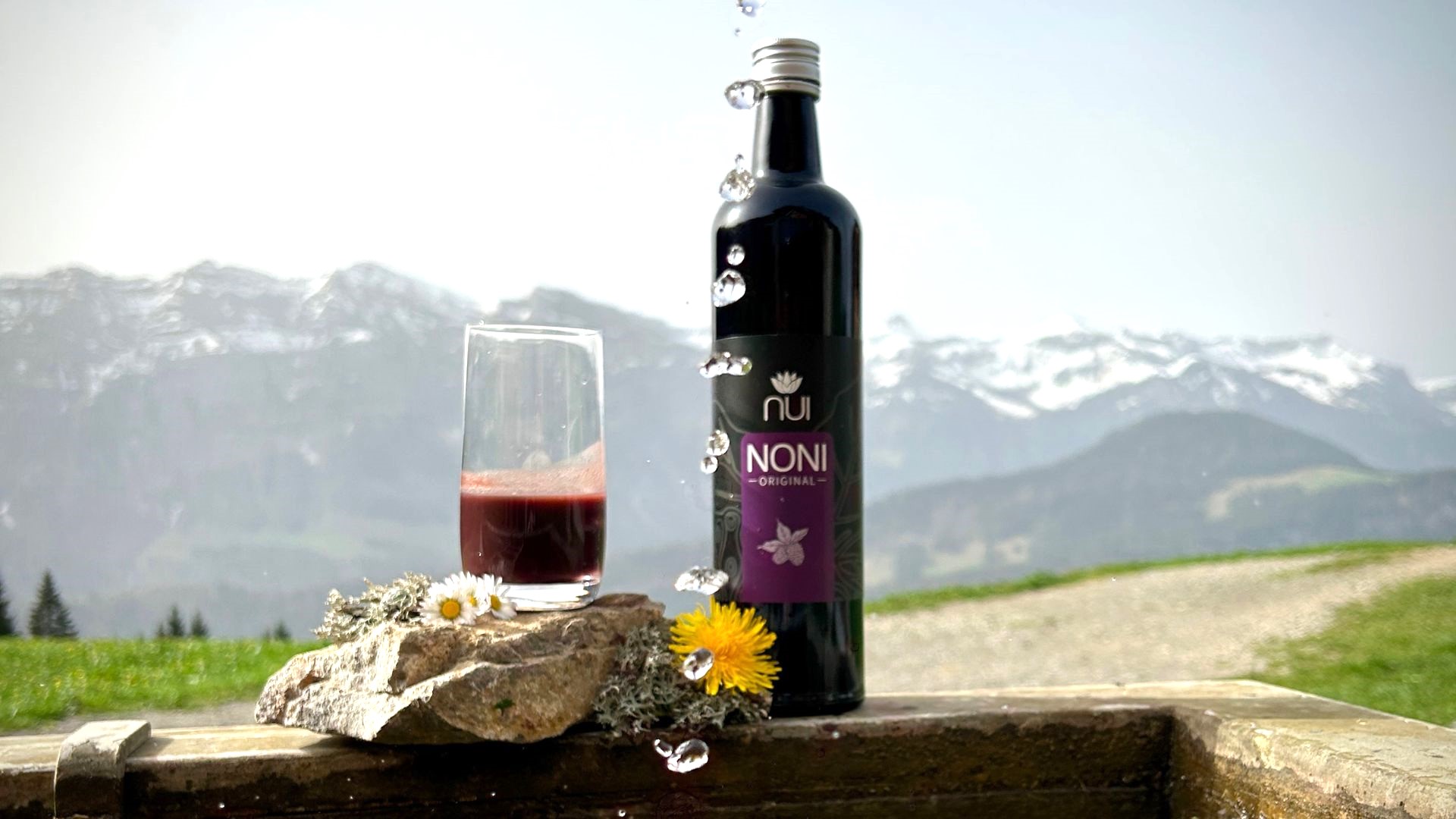 NUI NONI Original Flasche, daneben ein gefülltes Noni-Saftglas, steht an einem Brunnen im Hintergrund die Berge